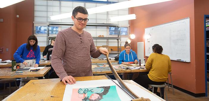 一个学生用一个大剪纸刀修剪他的艺术品，而其他学生在他身后的桌子上工作