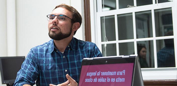 一个学生用西班牙语做报告，旁边是一台打开的笔记本电脑，上面用西班牙语显示报告的标题